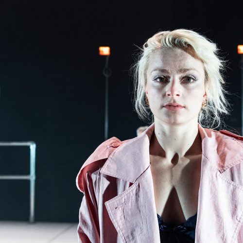 Young white blonde woman in a pink jacket standing on stage, looking into the camera