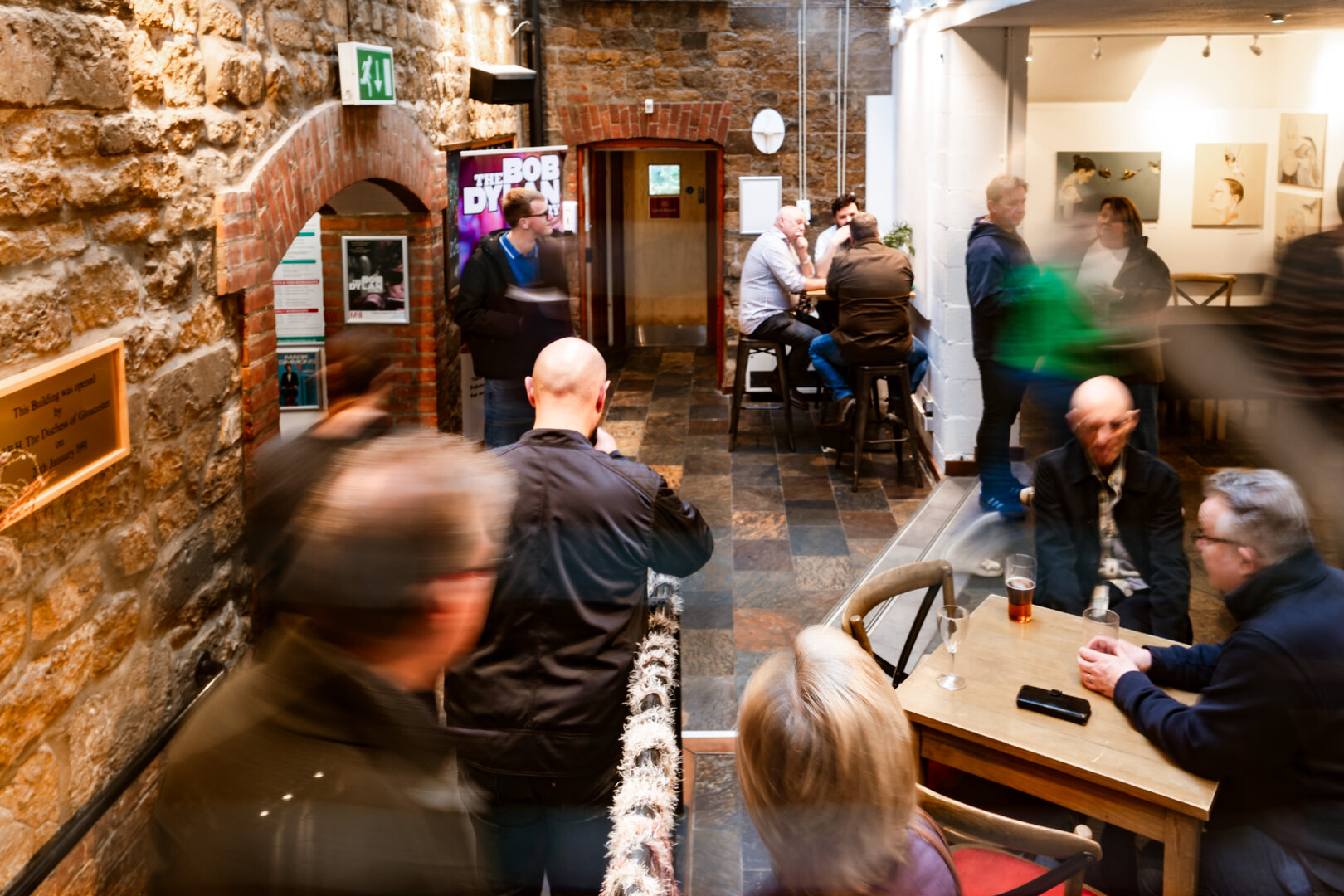 Group of people in the hallway of mill arts, there are some people sat on the right at a table and people walking through the doors