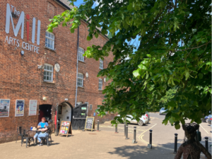 Exterior of The Mill Arts Centre with a tree in the middle, someone is sat outside at a table and chairs