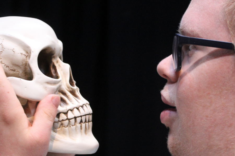 Person with short hair and glasses holding a skull prop up to their face