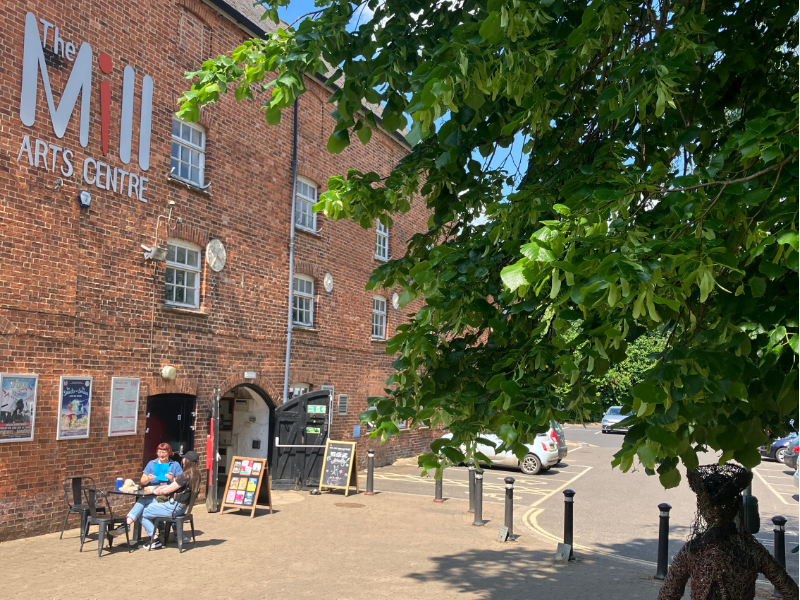Exterior of The Mill Arts Centre with a tree in the middle, someone is sat outside at a table and chairs